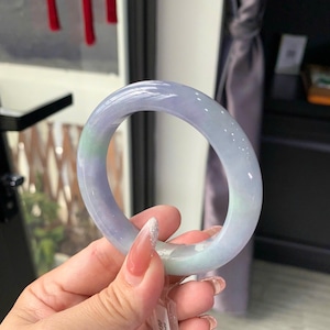 Op de afbeelding: Een gladde, cirkelvormige armband van lavendelkleurige jadeiet met subtiele doorschijnende eigenschappen en een klein lichtgroen kleurvlak. Een klein, doorzichtig plastic label met gedrukte tekst is aan de zijkant van de band bevestigd.