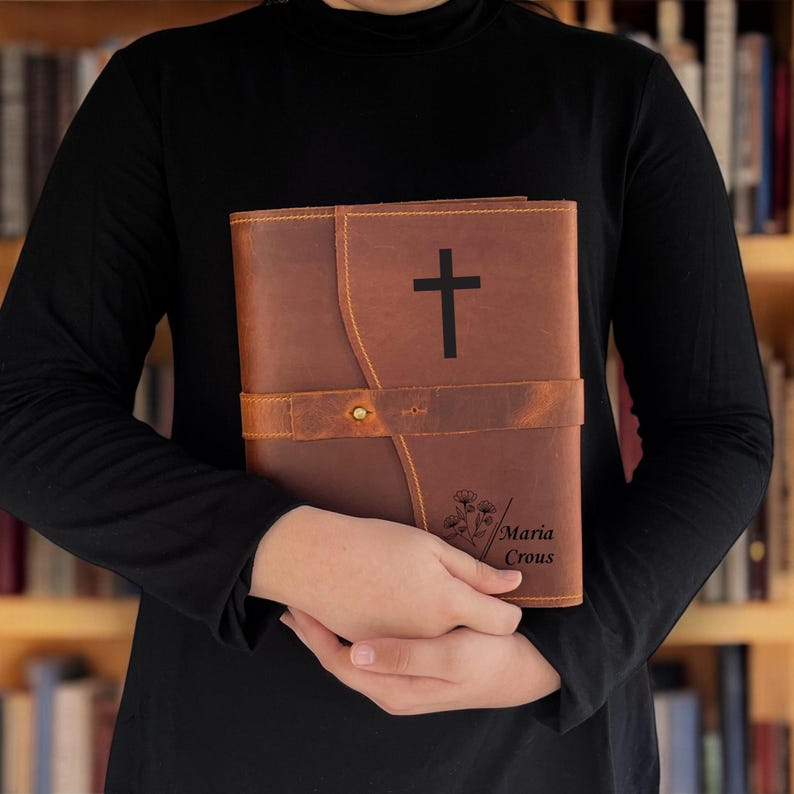 K&ouml;nnte beinhalten: Eine braune Lederbibel mit einem schwarzen Kreuz auf dem Einband. Die Bibel hat einen Riemenverschluss und der Name "Maria Crous" ist in der unteren rechten Ecke eingraviert. Die Bibel wird von einer Person gehalten, die ein schwarzes Langarmshirt tr&auml;gt.
