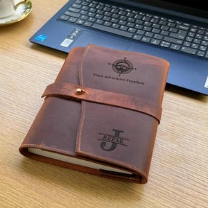 K&ouml;nnte beinhalten: Ein braunes Lederjournal mit Riemenverschluss und dem Text "Stars Adventure Freedom" und "Reese J" auf dem Einband eingraviert. Das Journal liegt offen auf einem Holztisch neben einem Laptop und einer Tasse Kaffee.