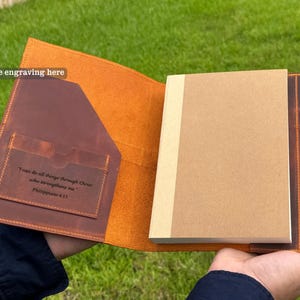K&ouml;nnte beinhalten: Offenes Lederjournal mit braunem Einband und einem beigefarbenen Notizblock. Das Journal hat einen Stifthalter mit einem goldenen Stift. Die Innenseite des Einbands hat eine Tasche und ein eingraviertes Zitat aus Philipper 4:13.