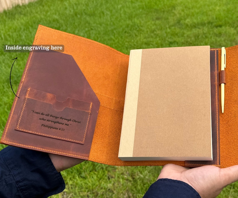 K&ouml;nnte beinhalten: Offenes Lederjournal mit braunem Einband und einem beigefarbenen Notizblock. Das Journal hat einen Stifthalter mit einem goldenen Stift. Die Innenseite des Einbands hat eine Tasche und ein eingraviertes Zitat aus Philipper 4:13.