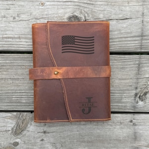 May include: A brown leather journal featuring a wrap-around strap with a brass stud closure. The front cover is engraved with a horizontal American flag graphic and a monogram consisting of the letter J with the name REESE centred across it.