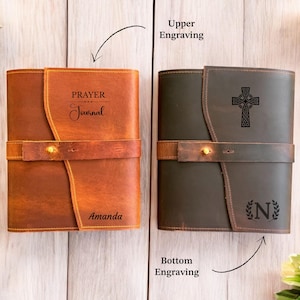 K&ouml;nnte beinhalten: Zwei Lederjournale, eines in Braun und eines in Dunkelgr&uuml;n, mit Riemenverschluss. Das braune Journal hat die Gravuren "PRAYER Journal" und "Amanda". Das gr&uuml;ne Journal zeigt ein keltisches Kreuz und den Buchstaben "N" mit Lorbeerkranz.