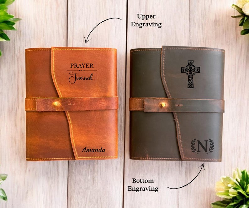 K&ouml;nnte beinhalten: Zwei Lederjournale, eines in Braun und eines in Dunkelgr&uuml;n, mit Riemenverschluss. Das braune Journal hat die Gravuren "PRAYER Journal" und "Amanda". Das gr&uuml;ne Journal zeigt ein keltisches Kreuz und den Buchstaben "N" mit Lorbeerkranz.