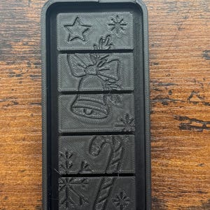 May include: Black rectangular mold with Christmas-themed designs. The mold features a star, a bell with a bow, a candy cane, and snowflakes. The mold is made of a dark material and is designed for crafting or baking.