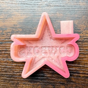 Puede incluir: Un molde rosa en forma de estrella con la palabra "COWBOYS" en una fuente de burbujas. El molde tiene un diseño en capas, con un interior rosa más claro y un contorno rosa más oscuro. El molde es probablemente para manualidades o repostería.