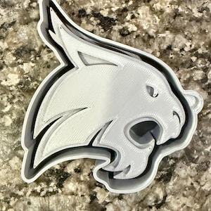 May include: A 3D-printed white and gray bobcat head with a detailed design. The bobcat is in profile, with its mouth open and a black outline. The design is layered, creating a three-dimensional effect. The object is likely a cookie cutter or a similar craft item.