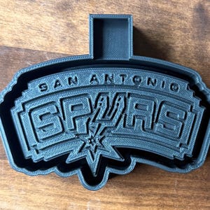 May include: A black 3D-printed cookie cutter in the shape of the San Antonio Spurs logo. The logo features the team name and a stylized spur and star design. The cutter has a handle for easy use.
