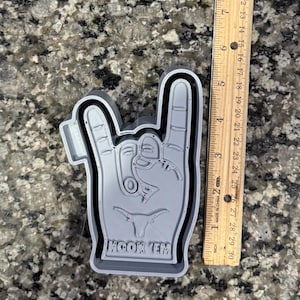 May include: A gray 3D printed cookie cutter shaped like a hand making the rock and roll hand gesture. The hand has a Longhorn steer head on the palm and the words "Hook 'em" at the bottom.