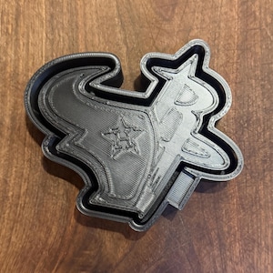 May include: A black plastic cookie cutter shaped like the Houston Texans logo. The logo features a star with the letter "H" inside, a bull's head, and a stylized "TX".