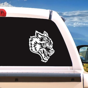 May include: White vinyl decal of a snarling wolf head, applied to the rear window of a vehicle. The wolf's detailed features include sharp teeth and textured fur, set against a black background.