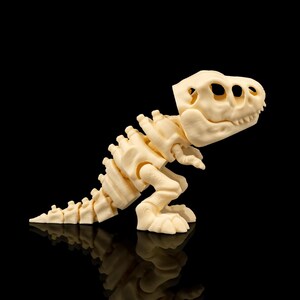 Puede incluir: Un juguete de esqueleto de dinosaurio T-Rex articulado de color crema. El juguete presenta un cráneo, columna vertebral y extremidades detallados, con articulaciones visibles para posar. El dinosaurio está sobre un fondo negro, creando un contraste llamativo.