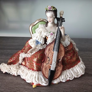 May include: A porcelain figurine of a person seated in a chair, playing a cello. The figure wears a red dress with gold patterns and white lace ruffles, featuring pink floral accents in the hair and on the bodice, with a green chair back.