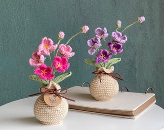 Könnte beinhalten: Zwei gehäkelte Blumenarrangements in beigen, kugelförmigen Töpfen. Ein Topf enthält rosa Blumen mit grünen Blättern, der andere violette Blumen mit grünen Blättern. Ein kleines braunes Etikett am rosa Blumentopf trägt die Aufschrift Thank you.