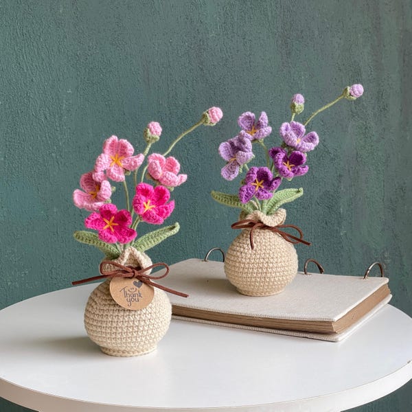 May include: Two crocheted flower arrangements in beige spherical pots. One pot holds pink flowers with green leaves, and the other holds purple flowers with green leaves. A small brown tag tied to the pink flower pot reads Thank you.