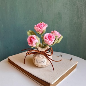 May include: A handmade crochet bouquet of pink roses with green leaves in a beige crocheted vase tied with a brown leather ribbon. The vase has a small tag that says "Handmade with love". The bouquet sits on a beige book.