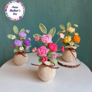 Könnte beinhalten: Drei handgefertigte gehäkelte Blumenarrangements in beigen Töpfen. Die Blumen sind in Rosa-, Lila-, Gelb- und Orangetönen mit grünen Blättern gehalten. Jeder Topf ist mit einem braunen Band gebunden. Das Bild enthält eine kreisförmige Grafik mit dem Text "Happy Mother's Day".