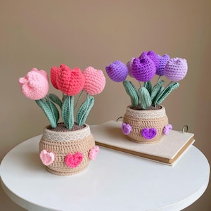 May include: Two crochet tulip flower arrangements in woven pots. One pot has pink and red tulips, the other has purple tulips. The pots are decorated with lace and heart accents. The flowers have green stems and leaves.