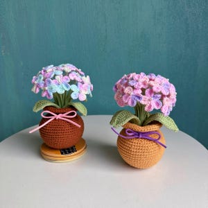 May include: Two handmade crochet potted flowers. One pot is brown with a pink bow, the other is orange with a purple bow. The flowers are pastel colors, with green leaves. The pots sit on a white surface.