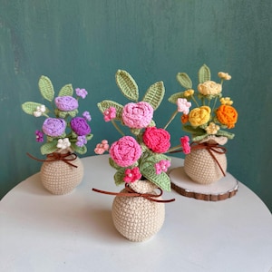 May include: Three crochet flower arrangements in small beige pots. The flowers are in shades of pink, purple, yellow, and orange, with green leaves. Each pot is tied with a brown ribbon. One arrangement sits on a small wooden slice.