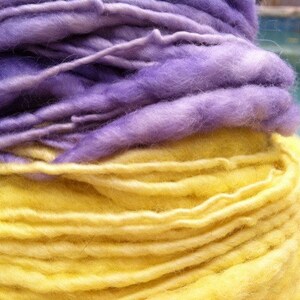 Puede incluir: Un ovillo de hilo de dos colores, un morado claro y un amarillo. El hilo está enrollado en forma de bola.