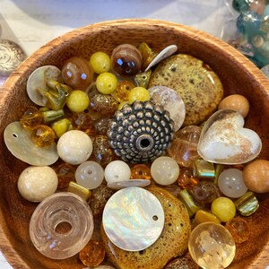 May include: A wooden bowl filled with a variety of beads in shades of yellow, brown, and white. The beads are made of different materials, including glass, wood, and shell.
