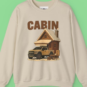 Pode incluir: Camisola bege com gola redonda com um gráfico de uma cabana em cima de uma carrinha Ford amarela. A palavra "CABIN" está impressa acima da cabana. A cabana tem uma chaminé e uma pequena mesa com duas cadeiras. O fundo é verde claro.