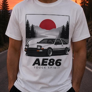 May include: A white t-shirt featuring a graphic print of a white and black AE86 car on a winding road. The design includes a large red sun, silhouetted pine trees, and the text AE86 TOUGE SPIRIT printed in bold black lettering below the car illustration.