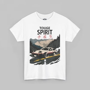 Puede incluir: Camiseta blanca con un gráfico de un coche deportivo blanco en una carretera de montaña. El diseño incluye las palabras "TOUGE SPIRIT" y caracteres japoneses. El coche tiene detalles en rojo y está ambientado en un paisaje de montañas y árboles.
