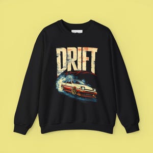 May include: Black sweatshirt featuring a graphic of a white and red sports car drifting, with the word "DRIFT" in large, distressed lettering above. The car is depicted with blue smoke and a sunset-like background.