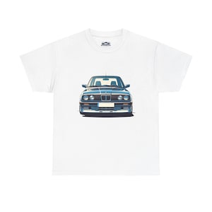 Puede incluir: Camiseta blanca con una ilustración frontal de un coche azul clásico. El coche tiene una parrilla delantera rectangular, faros redondos y un diseño cuadrado. La camiseta tiene un pequeño logotipo de coche en la parte superior del pecho.