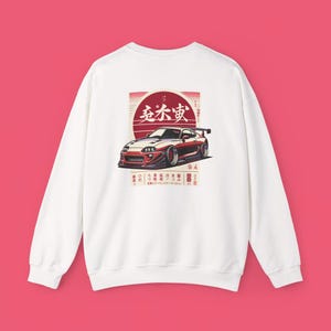 Op de afbeelding: Witte sweatshirt met een rode en zwarte afbeelding van een sportwagen met een spoiler, tegen een rode zon. Japanse tekst staat boven de auto. De sweatshirt heeft een ronde hals en lange mouwen.