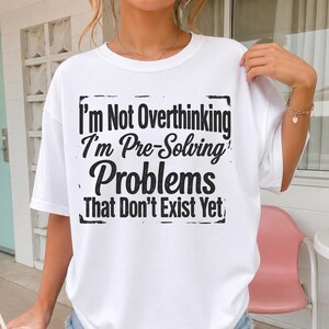May include: A white short-sleeved t-shirt featuring black text that reads, I'm Not Overthinking I'm Pre-Solving Problems That Don't Exist Yet, enclosed within a rectangular border with distressed edges on the front of the garment.