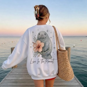 May include: A white long-sleeve sweatshirt featuring a graphic of a manatee next to a large peach-colored hibiscus flower, surrounded by small star icons. The text "Live In The Moment" is printed in black script below the manatee illustration.