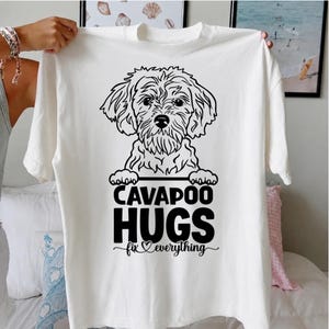 May include: A white short-sleeved t-shirt featuring a black line art illustration of a Cavapoo dog. Below the dog, the text reads CAVAPOO HUGS fix everything, with a small heart symbol integrated into the word fix in a cursive script font.
