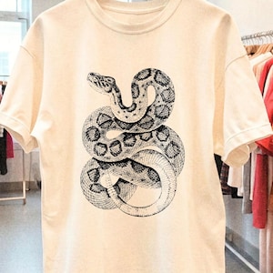 May include: Cream-colored t-shirt featuring a detailed black and white illustration of a coiled snake. The snake's scales and patterns are intricately depicted, creating a striking contrast against the shirt's neutral background. The shirt has short sleeves and a crew neck.