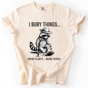 May include: Cream-colored t-shirt featuring a black and white illustration of a raccoon wearing overalls and a straw hat, digging with a small shovel. Text reads "I BURY THINGS... MAYBE PLANTS... MAYBE PEOPLE."