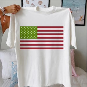 Könnte beinhalten: Ein weißes kurzärmeliges T-Shirt mit einem grafischen Flaggenaufdruck auf der Brust. Das Flaggendesign besteht aus einem grünen Feld mit weißen Sternen, das neben horizontalen roten und weißen Streifen angeordnet ist.