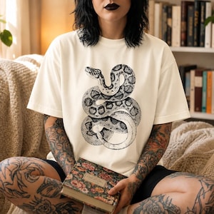 May include: An off-white short-sleeve t-shirt featuring a large, detailed black ink illustration of a coiled snake on the chest. The snake is depicted with intricate scale patterns and a winding, S-shaped body that occupies the center of the garment.