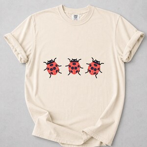 Pode incluir: Uma t-shirt de manga curta na cor creme com uma estampa gráfica de três joaninhas vermelhas com pintas pretas dispostas numa fila horizontal ao centro. A t-shirt tem um decote redondo e uma etiqueta da marca Comfort Colors na gola.