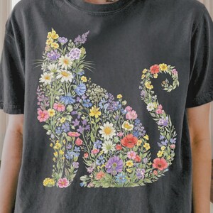 以下が含まれることがあります： 黒に近いグレーのTシャツ。花の猫のデザインが特徴です。猫のシルエットは、黄色、ピンク、紫、青の色合いのカラフルな花で埋められています。このシャツは半袖で、クルーネックです。