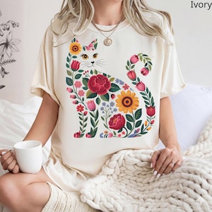Può includere: T-shirt color avorio con un design floreale a forma di gatto. Il gatto è composto da fiori colorati, tra cui rose rosse, girasoli gialli e tulipani rosa. Il design è centrato sulla maglietta, creando un aspetto stravagante e artistico. La maglietta ha maniche corte.