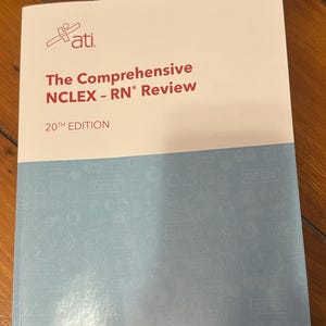 Puede incluir: Un libro de texto titulado "The Comprehensive NCLEX-RN Review" con el logotipo "ati". El libro de la 20ª edición tiene una cubierta blanca con detalles en azul y está sobre una superficie de madera.