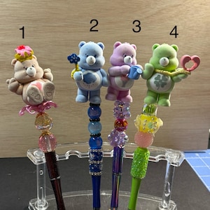 Peut inclure: Quatre stylos décoratifs avec des figurines de Bisounours. Chaque stylo a un personnage de Bisounours différent sur le dessus, avec des accents de perles colorées et un porte-stylo en acrylique transparent. Les stylos ont des corps de couleur foncée.