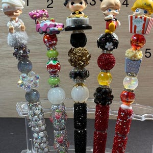 May include: Five decorative pens with various character toppers. The pens feature a variety of colors, including silver, red, black, and white. The pen designs include a figure with a white hat, a pink car, a black cat, Mickey Mouse, and a popcorn container.