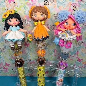 Könnte beinhalten: Drei dekorative Stifte mit modellierten Polymer-Ton-Figuren als Aufsatz. Stift 1 zeigt eine Puppe mit schwarzem Haar und Sonnenblumen. Stift 2 zeigt eine Puppe in einem orangefarbenen Kleid. Stift 3 zeigt eine Puppe mit rosa Haar und Pompons.