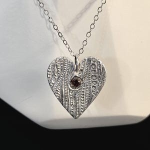 May include: A silver heart-shaped pendant necklace with a textured surface and a small, dark red gemstone in the center.