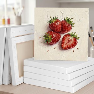 May include: A square canvas art print featuring a still life of strawberries. The painting shows three whole strawberries and one cut in half, with visible seeds. The canvas is stacked on top of other blank canvases, ready for painting.