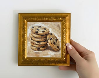 May include: A framed oil painting depicts a stack of six chocolate chip cookies, with one cookie leaning against the stack. The cookies are light brown with dark chocolate chips. The painting is in a gold ornate frame.