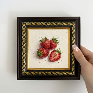 May include: A framed painting of strawberries. The artwork features four ripe, red strawberries with green leaves against a textured white background. The frame is dark brown with gold accents. The painting is held by a hand.